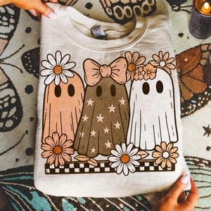 Op de afbeelding: Cr&egrave;mekleurige sweatshirt met een grillig ontwerp van drie spookfiguren versierd met bloemen en strikken. De geesten zijn in tinten perzik, bruin en wit, met ster- en madeliefjesaccenten. Een zwart-wit geblokte rand maakt het ontwerp compleet.