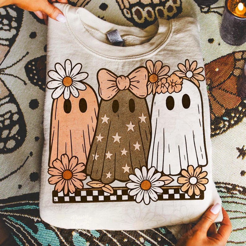 Op de afbeelding: Cr&egrave;mekleurige sweatshirt met een grillig ontwerp van drie spookfiguren versierd met bloemen en strikken. De geesten zijn in tinten perzik, bruin en wit, met ster- en madeliefjesaccenten. Een zwart-wit geblokte rand maakt het ontwerp compleet.