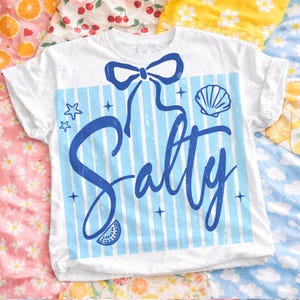 May include: White t-shirt with a blue bow, blue and white striped background, and the word "Salty" in a script font. The design includes a seashell, stars, and a watermelon slice.