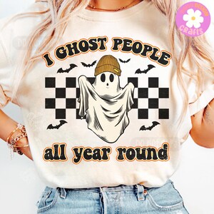 May include: Cream-coloured t-shirt featuring a ghost wearing a beanie and the text "I ghost people all year round." The design includes black bats and a checkered pattern, suitable for Halloween or general wear.