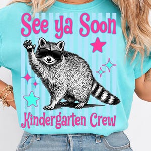 Op de afbeelding: Een lichtblauw T-shirt met de roze tekst "See ya Soon Kindergarten Crew". Het shirt heeft een wasbeer met een zonnebril die zwaait. Roze en turquoise sterren en lijnen versieren de achtergrond.