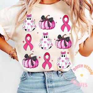 May include: A white t-shirt with a pink ribbon, ghosts, and pumpkins. The design features a pink ribbon, white ghosts with pink flowers, and pink pumpkins with black polka dots and black bows. The text "Daisy Design Crafts" is visible in the bottom right corner.