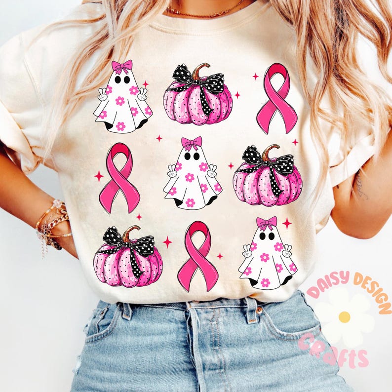 May include: A white t-shirt with a pink ribbon, ghosts, and pumpkins. The design features a pink ribbon, white ghosts with pink flowers, and pink pumpkins with black polka dots and black bows. The text "Daisy Design Crafts" is visible in the bottom right corner.