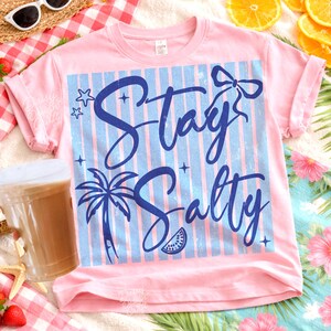 May include: A light pink t-shirt with rolled-up sleeves. The shirt features a blue and white striped design with the words "Stay Salty" in a blue script font, along with a palm tree, bow, stars, and a watermelon slice.