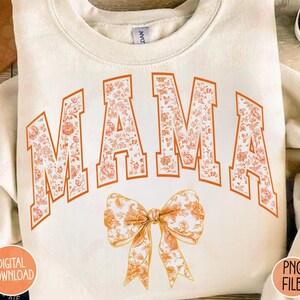 May include: A white sweatshirt with the word "MAMA" in orange letters with a floral pattern. A large orange bow with a floral pattern is below the word.