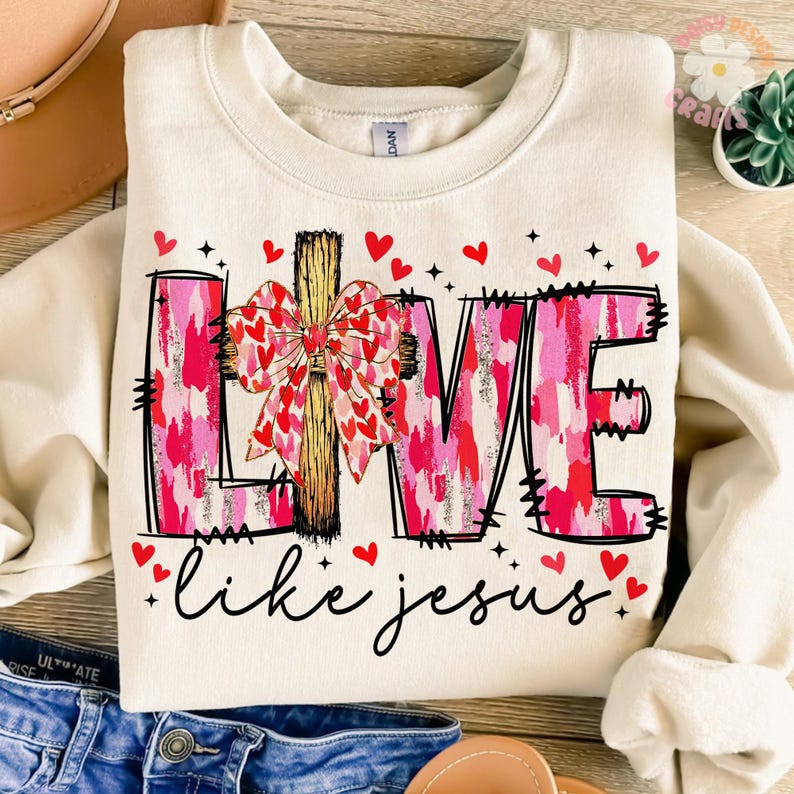 May include: A cream-coloured sweatshirt with a pink and white camouflage pattern that spells out "LOVE" with a cross and bow in the centre. The text "like jesus" is written below the word "LOVE" in black.