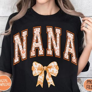 May include: Black t-shirt with the word "NANA" in orange letters with a floral pattern. The word is in a collegiate font style. A floral bow is below the word.