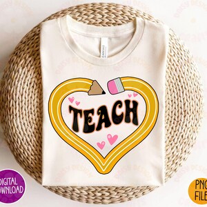 K&ouml;nnte beinhalten: Ein wei&szlig;es T-Shirt mit einem gelben Bleistiftdesign in Form eines Herzens. Das Wort "TEACH" ist in schwarzen Buchstaben innerhalb des Herzens geschrieben. Um das Bleistiftdesign herum sind rosa Herzen verstreut.