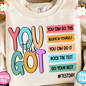 May include: A beige sweatshirt with the words "You Got This" in colorful letters. The words "You Can Do This", "Believe In Yourself", "You Can Do It", "Rock The Test", and "Do Your Best" are written in colorful stripes below the main text. The hashtag "#TestDay" is written in black at the bottom of the sweatshirt.