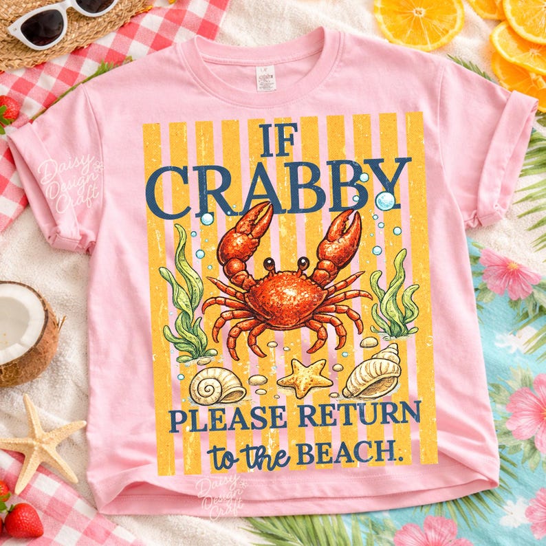 May include: Pink t-shirt with a crab graphic and the text "IF CRABBY PLEASE RETURN to the BEACH." The design includes a red crab, seashells, and seaweed against a yellow striped background. The shirt is short-sleeved.