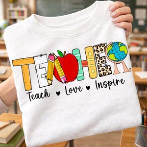Puede incluir: Una camiseta blanca con la palabra TEACHER en letras coloridas con tem&aacute;tica escolar, incluyendo una regla, un l&aacute;piz, una manzana roja, libros y un globo terr&aacute;queo. Debajo, el texto Teach Love Inspire aparece en fuente negra con corazones.