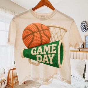 K&ouml;nnte beinhalten: Cremefarbenes T-Shirt mit Basketball-, Netz- und Megaphon-Grafik. Der Basketball ist orange und braun, das Netz beige und das Megaphon gr&uuml;n mit wei&szlig;em Text "GAME DAY". Das Shirt h&auml;ngt an einem Holzb&uuml;gel.
