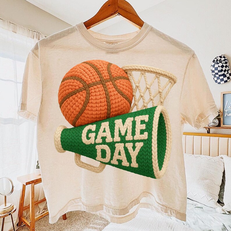 K&ouml;nnte beinhalten: Cremefarbenes T-Shirt mit Basketball-, Netz- und Megaphon-Grafik. Der Basketball ist orange und braun, das Netz beige und das Megaphon gr&uuml;n mit wei&szlig;em Text "GAME DAY". Das Shirt h&auml;ngt an einem Holzb&uuml;gel.