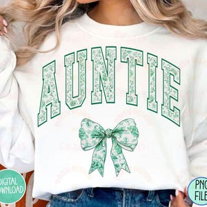 May include: White sweatshirt with the word "AUNTIE" in green letters with a floral pattern. A green floral bow is below the text.
