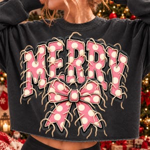 May include: A charcoal grey long-sleeve sweatshirt featuring the word MERRY in large pink letters with white polka dots. The letters and a large pink polka-dot bow below are outlined with frayed, light-coloured decorative stitching.