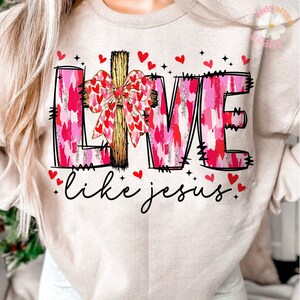 May include: Beige sweatshirt with the word "LOVE" in pink and red brushstroke letters, a wooden cross, and a bow made of hearts. The phrase "like jesus" is written in cursive below, with red heart accents.