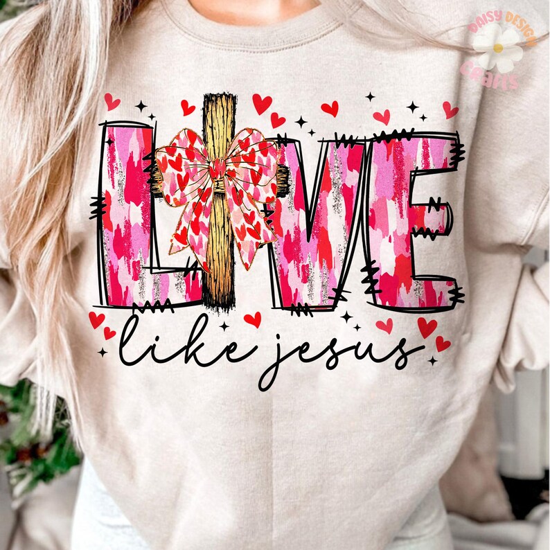 May include: Beige sweatshirt with the word "LOVE" in pink and red brushstroke letters, a wooden cross, and a bow made of hearts. The phrase "like jesus" is written in cursive below, with red heart accents.