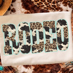 Pu&ograve; includere: Una maglietta beige con la scritta "RODEO" in grandi lettere consumate. Le lettere sono riempite con un motivo a stampa di pelle di mucca e leopardo, delineate in azzurro. La maglietta ha un tema western.