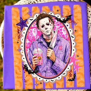 May include: A purple t-shirt featuring a graphic of a character with a white mask holding a knife and a pink drink with a pumpkin face. The design is framed by a scalloped oval border with bats and stars against a purple and orange striped background.