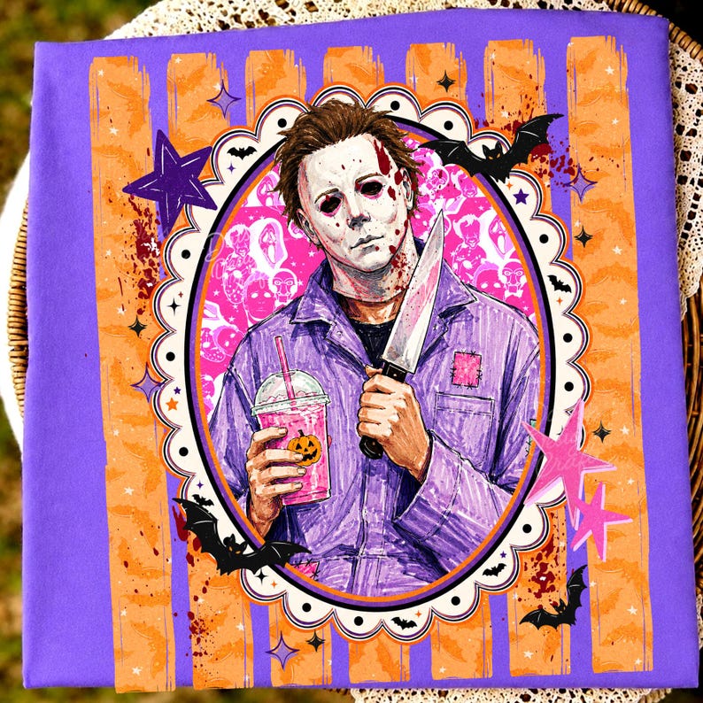 May include: A purple t-shirt featuring a graphic of a character with a white mask holding a knife and a pink drink with a pumpkin face. The design is framed by a scalloped oval border with bats and stars against a purple and orange striped background.