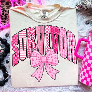 May include: Cream-colored t-shirt with the word "SURVIVOR" in pink, patterned letters. A pink bow with breast cancer awareness ribbons is below the word. The shirt has a crew neck and short sleeves.