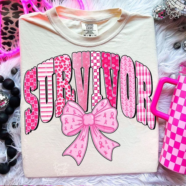 May include: Cream-colored t-shirt with the word "SURVIVOR" in pink, patterned letters. A pink bow with breast cancer awareness ribbons is below the word. The shirt has a crew neck and short sleeves.