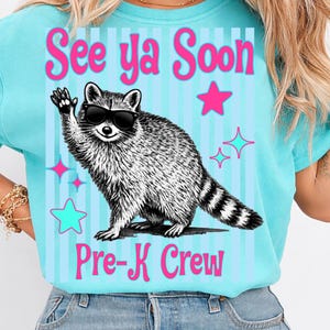 Op de afbeelding: Een lichtblauw T-shirt met de tekst "See ya Soon Pre-K Crew". Het shirt heeft een cartoon wasbeer met een zonnebril die zwaait. Roze sterren en blauwe sterrenhemels versieren het shirt.