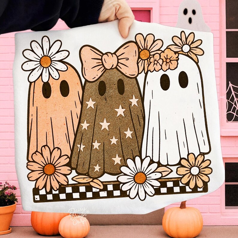 May include: A white tote bag featuring three cartoon ghosts with daisy accents. One ghost has a bow, another has star patterns, and the third has a daisy crown. The design includes a black and white checkered border and is decorated with orange and white daisies.