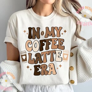Puede incluir: Camiseta blanca con texto marr&oacute;n y beige que dice "In My Coffee Latte Era" con dos ilustraciones de tazas de caf&eacute;.