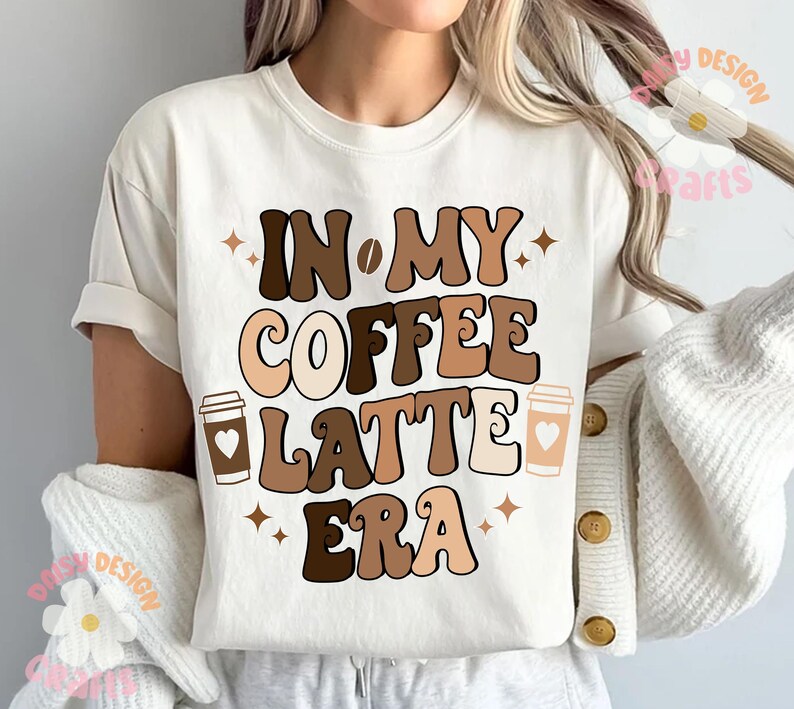 Puede incluir: Camiseta blanca con texto marr&oacute;n y beige que dice "In My Coffee Latte Era" con dos ilustraciones de tazas de caf&eacute;.