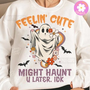 May include: White sweatshirt featuring a ghost design with flowers and a rainbow. The text reads "FEELIN' CUTE" and "MIGHT HAUNT U LATER. IDK". The design includes black bats and stars, creating a fun, spooky aesthetic.