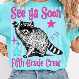 Op de afbeelding: Een lichtblauw T-shirt met de tekst "See ya Soon Fifth Grade Crew" in roze. Het shirt heeft een wasbeer met een zonnebril die zwaait, met roze en turquoise sterren en strepen.