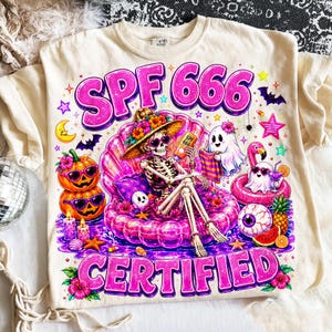 May include: A cream-colored t-shirt featuring a graphic of a skeleton wearing a floral hat and heart-shaped sunglasses, lounging in a pink inflatable shell. The text reads SPF 666 CERTIFIED. Includes pumpkins, ghosts, and tropical elements.