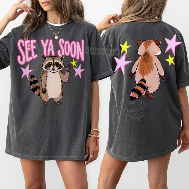 K&ouml;nnte beinhalten: Dunkelgraues T-Shirt mit einem Waschb&auml;r-Motiv und dem Text "SEE YA SOON" in Pink. Der Waschb&auml;r ist braun, hellbraun und schwarz, mit rosa und gelben Sternen. Auf der R&uuml;ckseite des Shirts sind ein Waschb&auml;r und Sterne abgebildet.