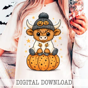 May include: A cream-colored t-shirt featuring a cartoon Highland cow wearing a gray beanie with pumpkin faces, sitting on an orange pumpkin. The cow is wearing a white outfit with pumpkin buttons. The text "DIGITAL DOWNLOAD" is at the bottom.
