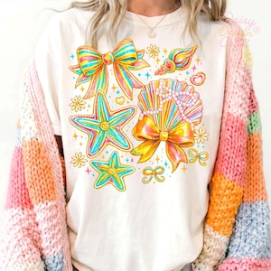 May include: White t-shirt featuring colorful illustrations of bows, starfish, seashells, and flowers. The design includes a large bow, two starfish, a seashell with pearls, and several smaller bows and flowers. The shirt is paired with a multi-colored sweater.