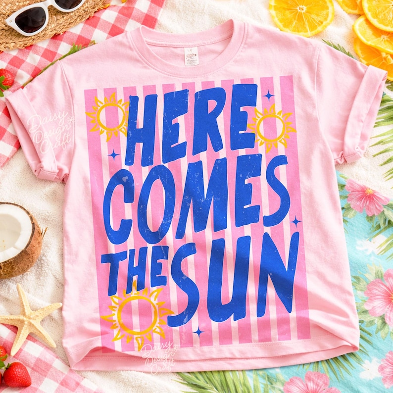 May include: Pink t-shirt with the text "HERE COMES THE SUN" in blue, set against a striped background. Yellow sun graphics are placed around the text. The shirt is a casual wear item.