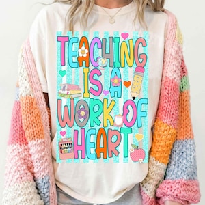 Puede incluir: Una camiseta blanca con un gr&aacute;fico colorido que incluye el texto "TEACHING IS A WORK OF HEART" en letras grandes y multicolores. El dise&ntilde;o presenta ilustraciones de una pila de libros, un l&aacute;piz, una caja de ceras y una manzana.
