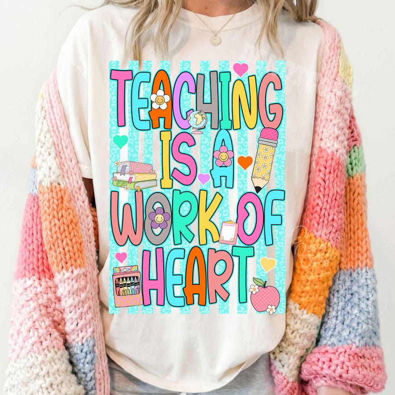 Puede incluir: Una camiseta blanca con un gr&aacute;fico colorido que incluye el texto "TEACHING IS A WORK OF HEART" en letras grandes y multicolores. El dise&ntilde;o presenta ilustraciones de una pila de libros, un l&aacute;piz, una caja de ceras y una manzana.