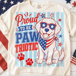 May include: Cream-colored t-shirt with a patriotic dog graphic. The design features a white dog wearing star-shaped sunglasses and a bandana with an American flag design. Text reads "Proud to be PAW TRIOTIC" in red and blue.