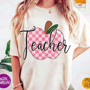 K&ouml;nnte beinhalten: Ein wei&szlig;es T-Shirt mit einem pink-wei&szlig; karierten Apfel-Design und dem Wort "Teacher" in schwarzer Schrift.