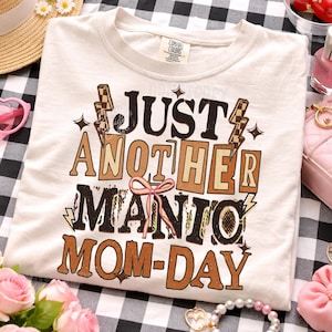 May include: Cream-colored t-shirt with the text "JUST ANOTHER MANIC MOM-DAY" in brown and tan lettering. The design includes lightning bolts and a pink bow. The shirt is on a black and white checkered surface.