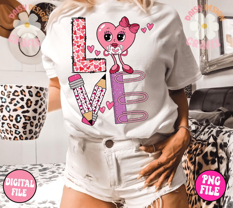 May include: White t-shirt featuring a pink heart design that reads "LOVE" in pink letters. The heart has a pink bow and large eyes. There are pink pencils and paper clips on either side of the heart.