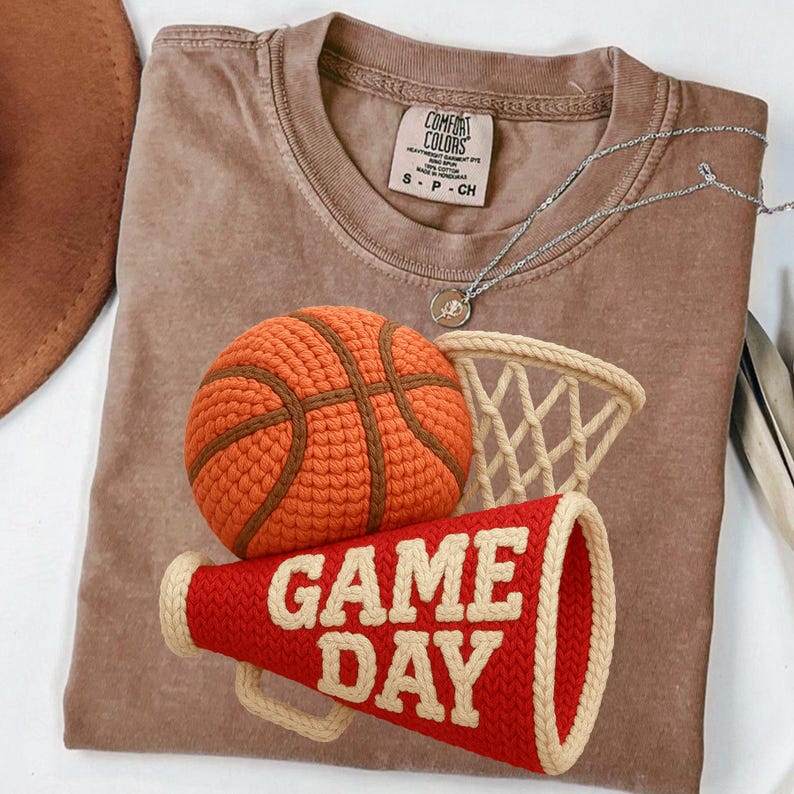 K&ouml;nnte beinhalten: Ein braunes T-Shirt mit einem Basketball-, Netz- und Megaphon-Design. Der Basketball ist orange mit braunen Linien, das Netz ist beige und das Megaphon ist rot mit "GAME DAY" in Wei&szlig;. Eine silberne Halskette ist ebenfalls sichtbar.