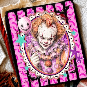 May include: A black t-shirt featuring a graphic print of a clown with orange hair, a ruffled collar, and a pink balloon. The design is framed by a scalloped oval border against a background of black vertical stripes, pink bats, and stars.