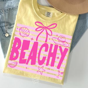May include: Yellow t-shirt with a pink graphic that says "BEACHY". The graphic includes a seashell, bow, hat, watermelon slice, star, and cocktail. The shirt is folded on top of blue jeans and a tan hat.