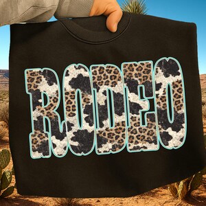 Pu&ograve; includere: Felpa nera con la scritta "RODEO" in grandi lettere sbiadite. Le lettere sono riempite con un motivo a pelle di mucca e leopardo, bordate di azzurro. Lo sfondo mostra un paesaggio desertico.