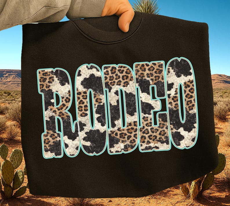 Pu&ograve; includere: Felpa nera con la scritta "RODEO" in grandi lettere sbiadite. Le lettere sono riempite con un motivo a pelle di mucca e leopardo, bordate di azzurro. Lo sfondo mostra un paesaggio desertico.