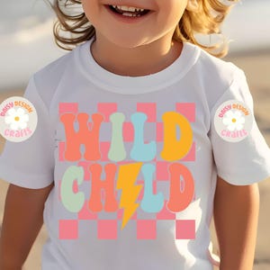 Op de afbeelding: Wit T-shirt met een roze en witte geruite achtergrond. De woorden "WILD CHILD" staan in kleurrijke retro-lettertypen met een gele bliksem ertussen.