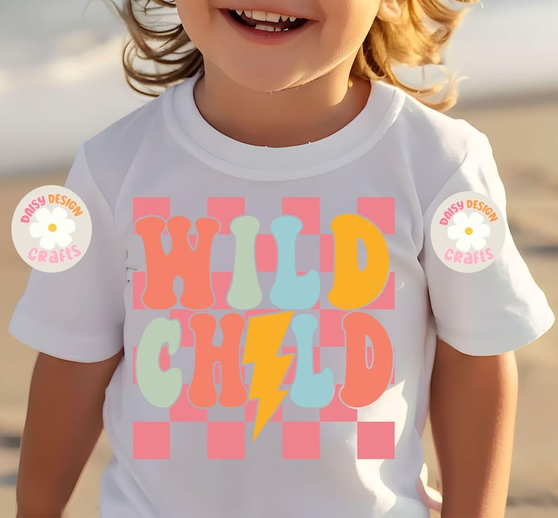 Op de afbeelding: Wit T-shirt met een roze en witte geruite achtergrond. De woorden "WILD CHILD" staan in kleurrijke retro-lettertypen met een gele bliksem ertussen.